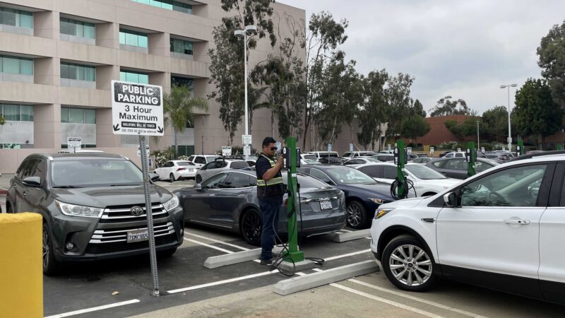 A person uses an electric vehicle charging station in a parking lot filled with cars, promoting clean transportation.