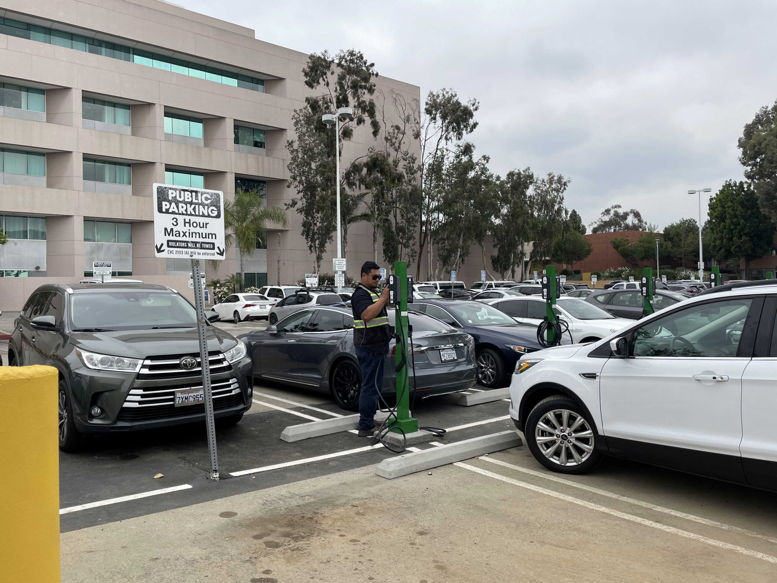 A person uses an electric vehicle charging station in a parking lot filled with cars, promoting clean transportation.