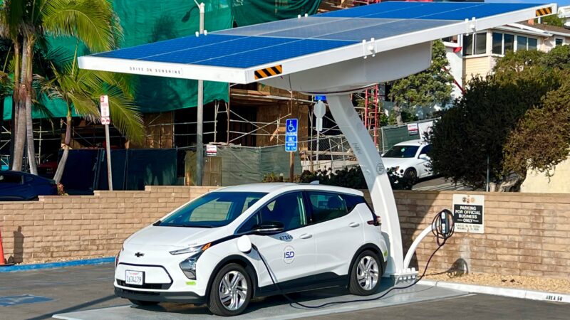 A white electric vehicle charges under a solar panel at a clean energy station in Los Angeles.
