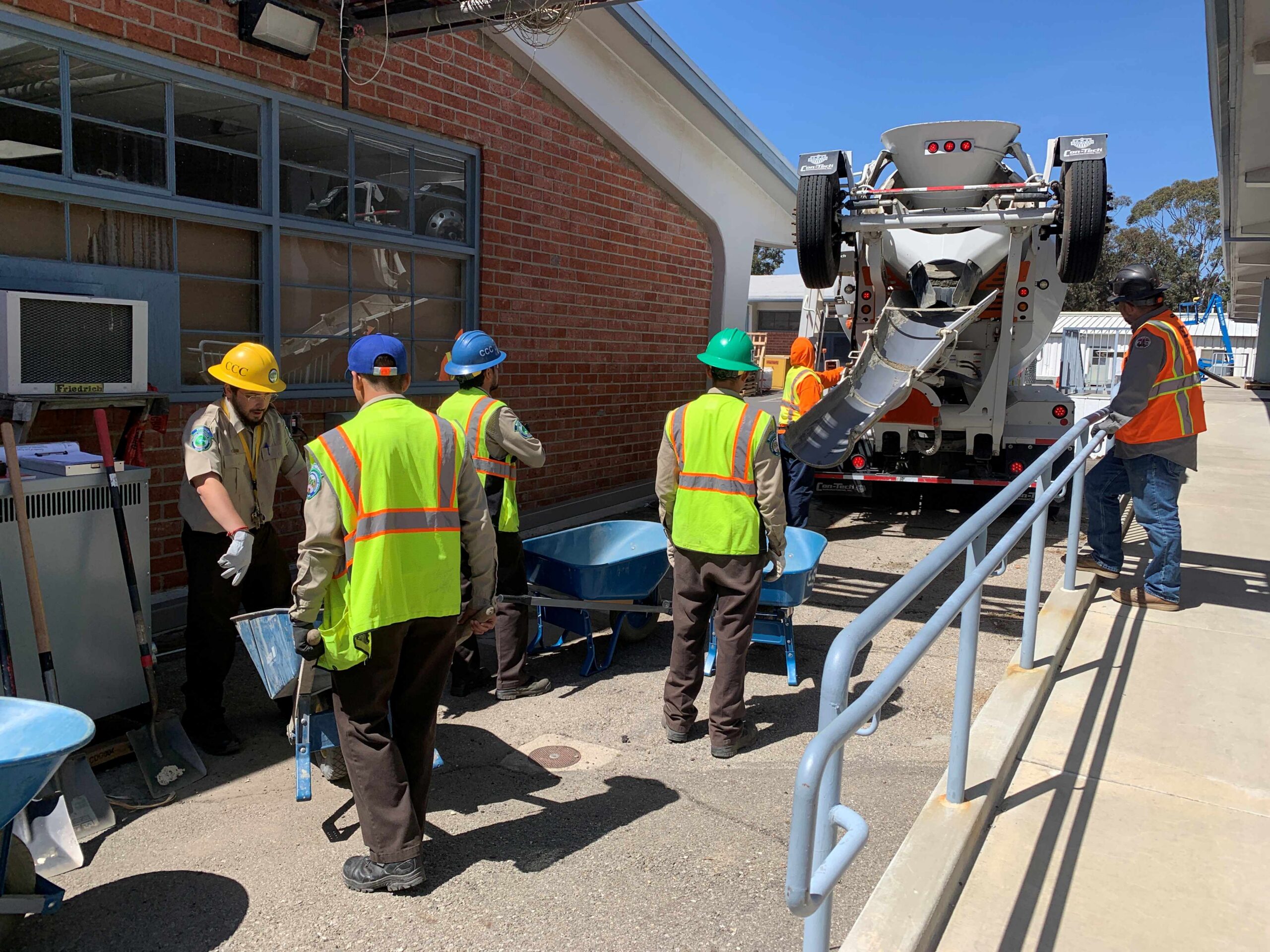 Workers in hard hats and safety vests operate equipment and wheelbarrows during a clean energy project at a facility.