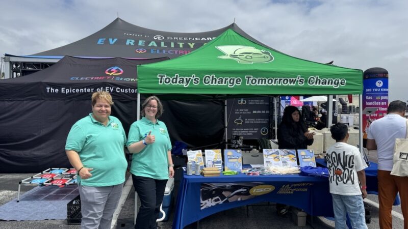 Two representatives stand at a booth promoting electric vehicles under a green tent at a community event.