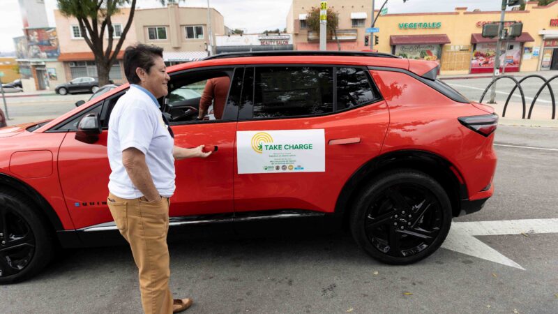 A participant approaches a red electric vehicle with a "Take Charge" sign during the clean mobility event in East Los Angeles.