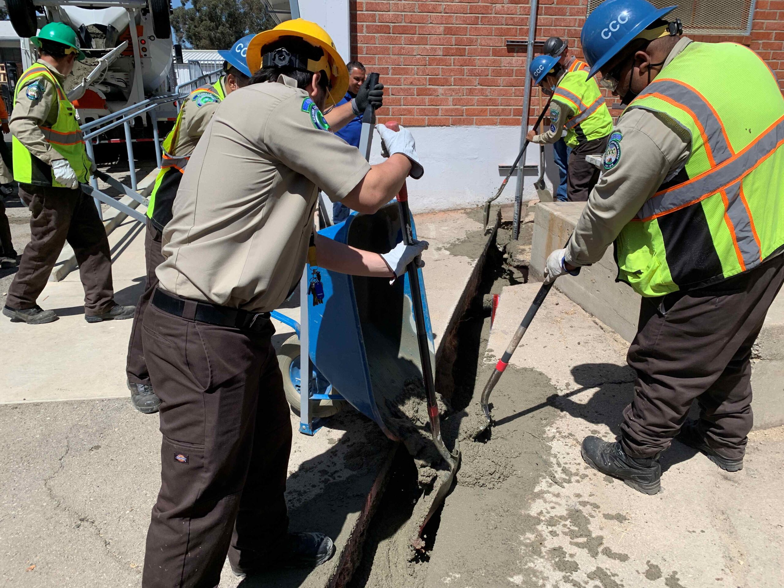 Workers wearing safety gear and hard hats pour and spread fresh concrete into a long trench beside a brick building, using shovels and a wheelbarrow to fill the area.