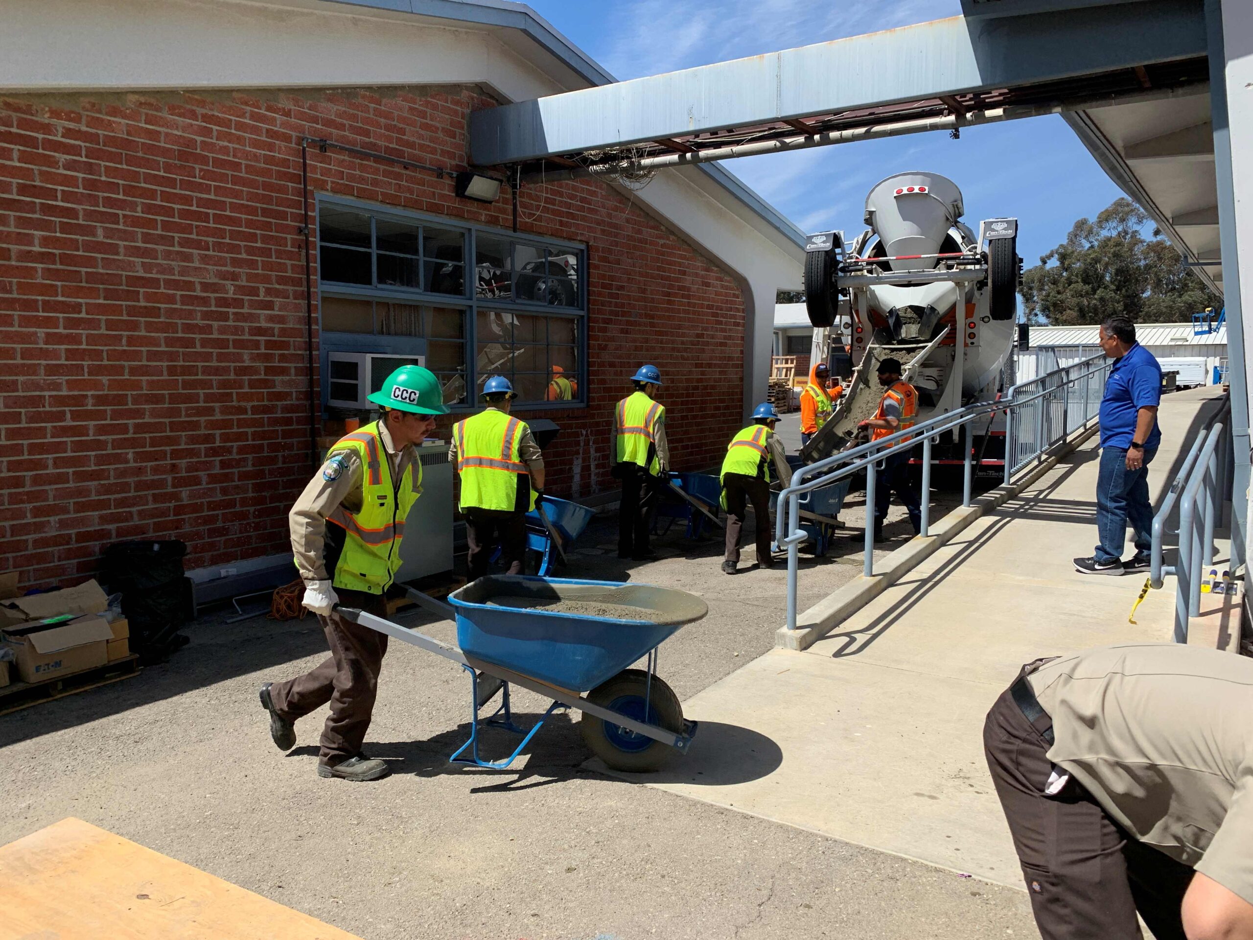 Workers in safety vests and hard hats transport concrete in wheelbarrows from a mixing truck, moving along a ramp beside a brick building as part of a construction project.
