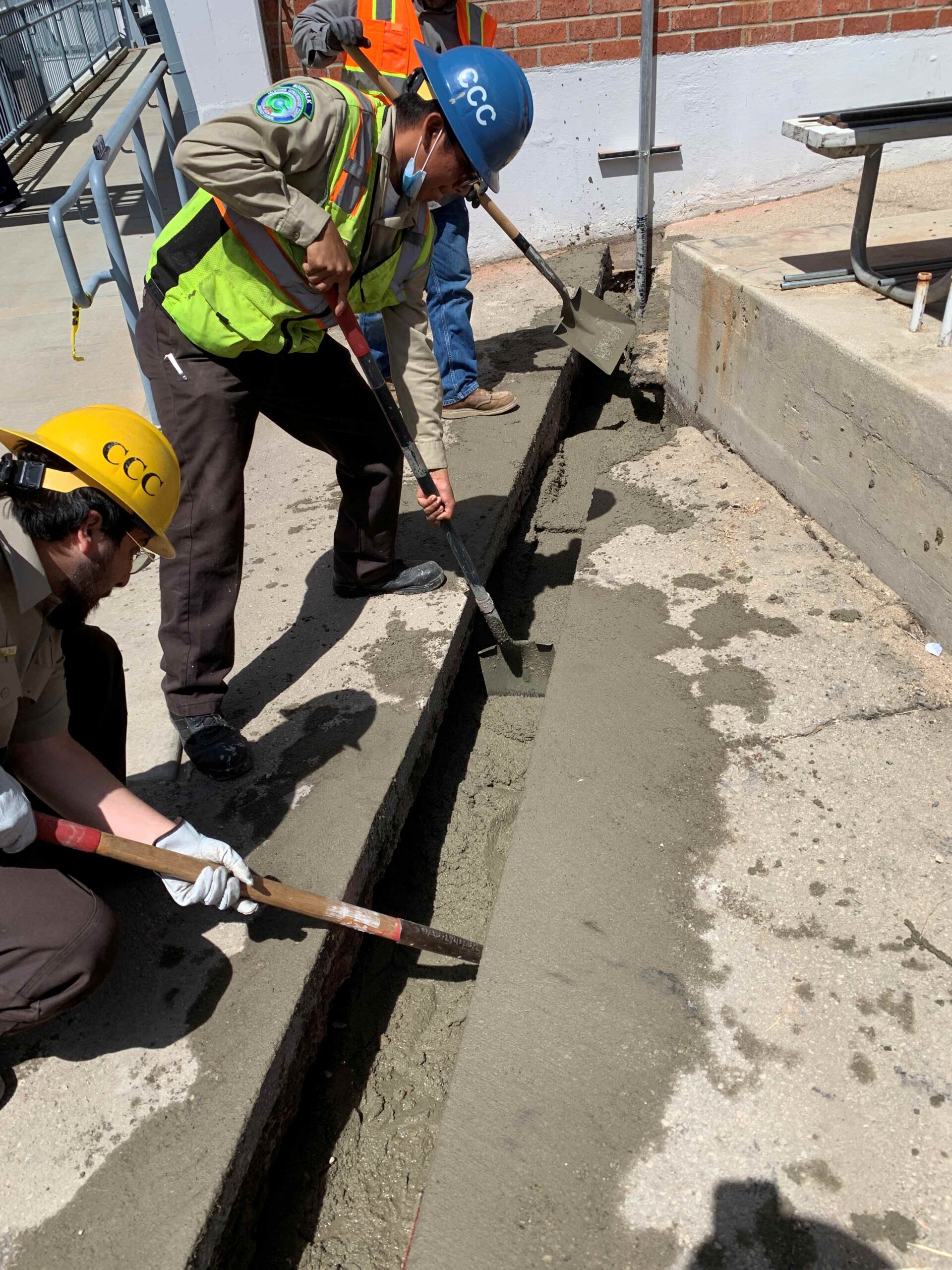 Workers wearing hard hats and safety vests spread and level wet concrete inside a long, narrow trench next to a brick building.