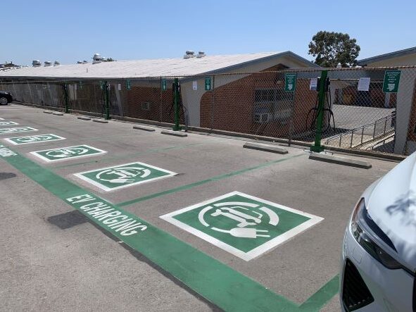 A row of newly marked EV charging parking spaces with green painted symbols sits next to a fenced area and charging stations, with a parked vehicle partially visible in the foreground.