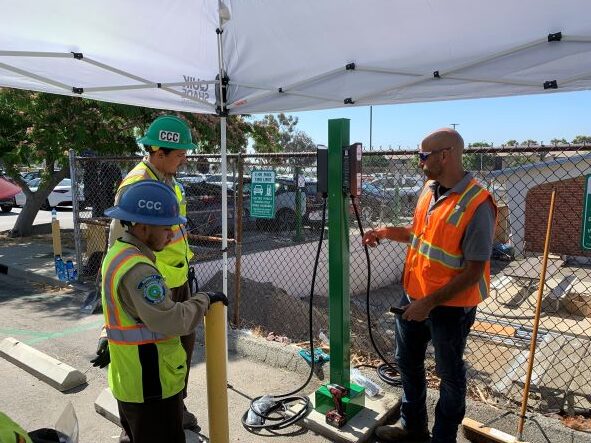 Two workers wearing safety vests and hard hats labeled &ldquo;CCC&rdquo; watch as an instructor demonstrates how to operate an electric vehicle charging station under a white canopy at an outdoor worksite.