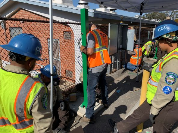 Workers in safety vests and hard hats gather around a newly installed green EV charging post, while a technician connects wiring at an outdoor worksite.