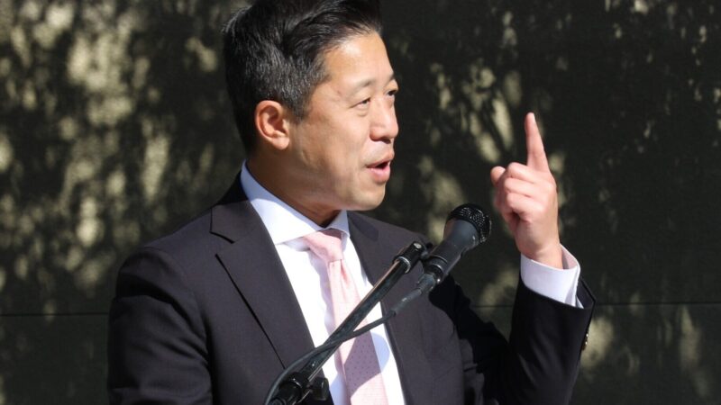 Michael Owh speaks at an outdoor podium, gesturing with one hand while addressing an audience through a microphone during a public event.