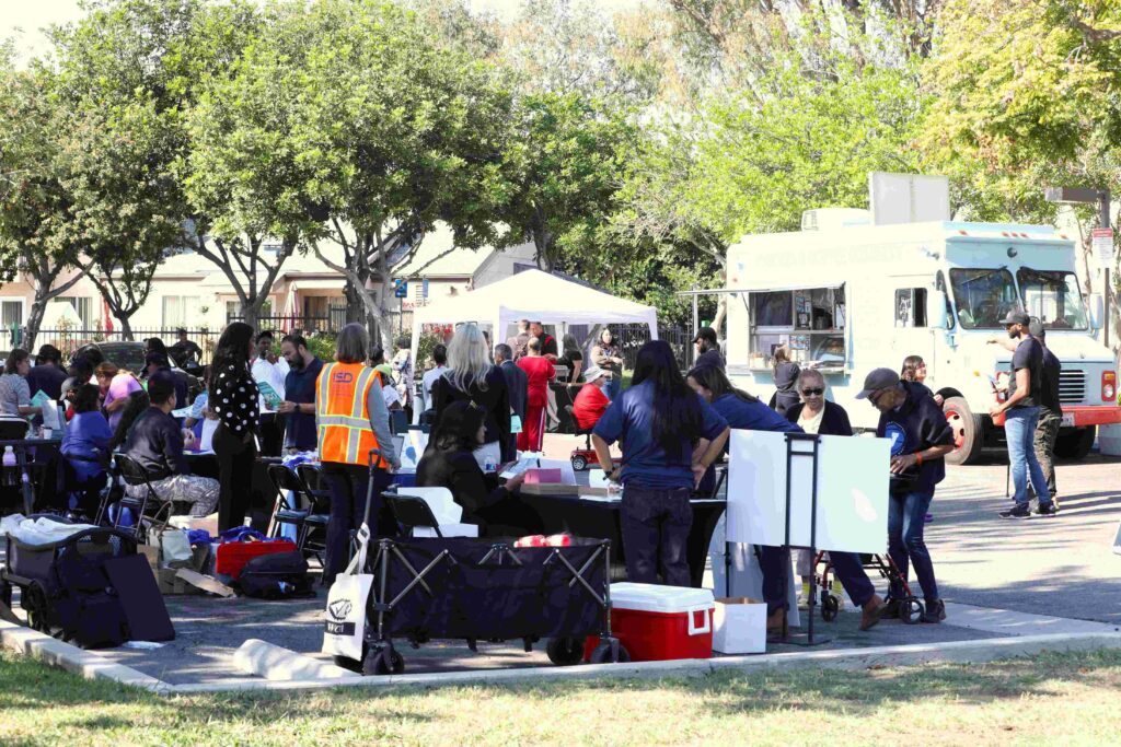 Community members gather at an outdoor resource fair with tables, tents, and service providers, as people receive assistance and information near a parked food truck in a park-like setting.