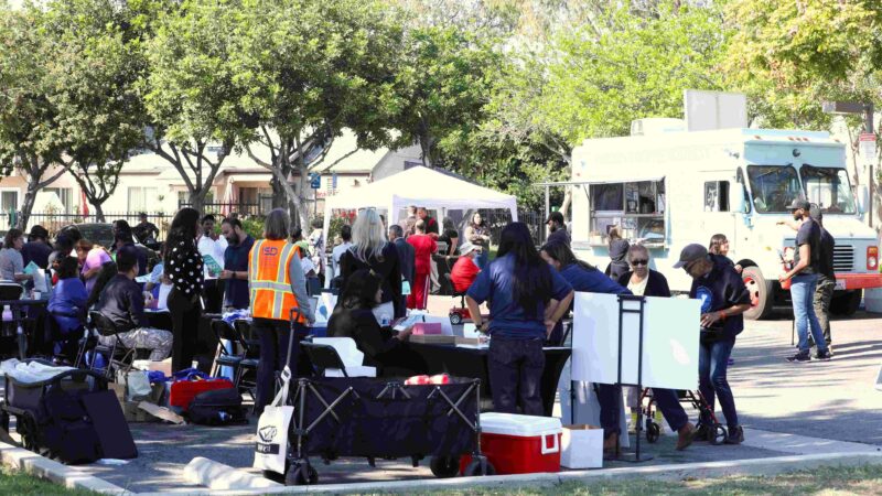 Community members gather at an outdoor resource fair with tables, tents, and service providers, as people receive assistance and information near a parked food truck in a park-like setting.