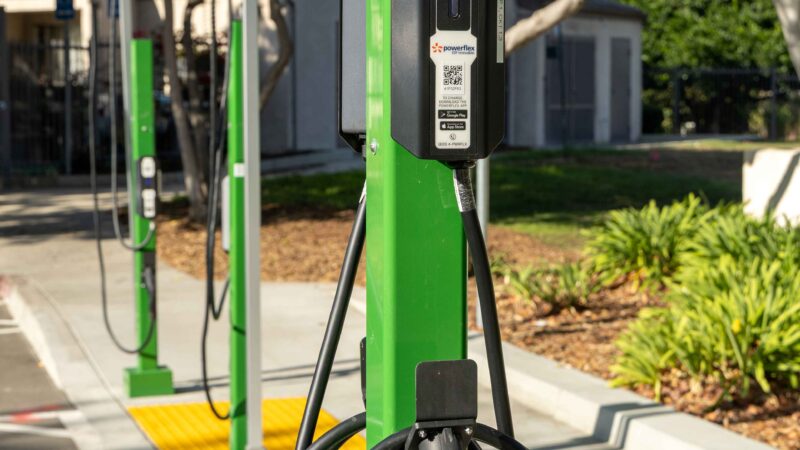 A row of green electric vehicle charging stations with coiled charging cables stands along a paved parking area, surrounded by trees and landscaping in an outdoor setting.