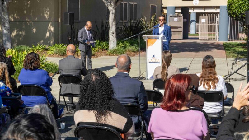 A speaker stands at a podium with an LACDA logo, addressing an audience seated outdoors during a daytime event, while another person stands nearby reading notes in front of a residential building.