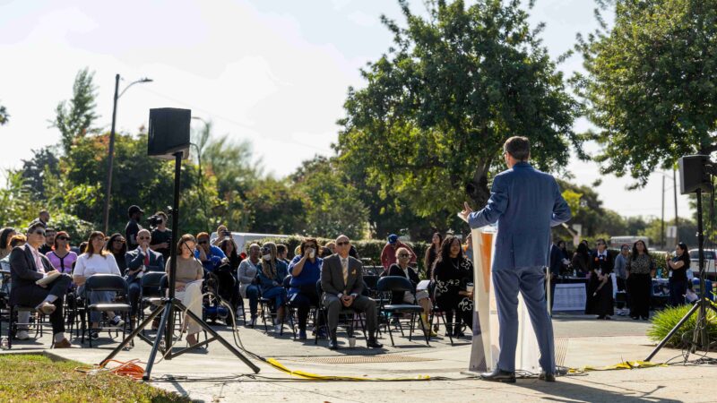 A speaker stands at a podium addressing a seated audience at an outdoor community event, with rows of chairs, speakers, and attendees gathered in a park-like setting on a sunny day.